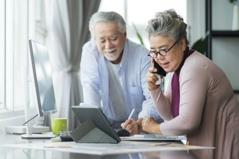 An older couple talking on the phone while sitting at a desk.