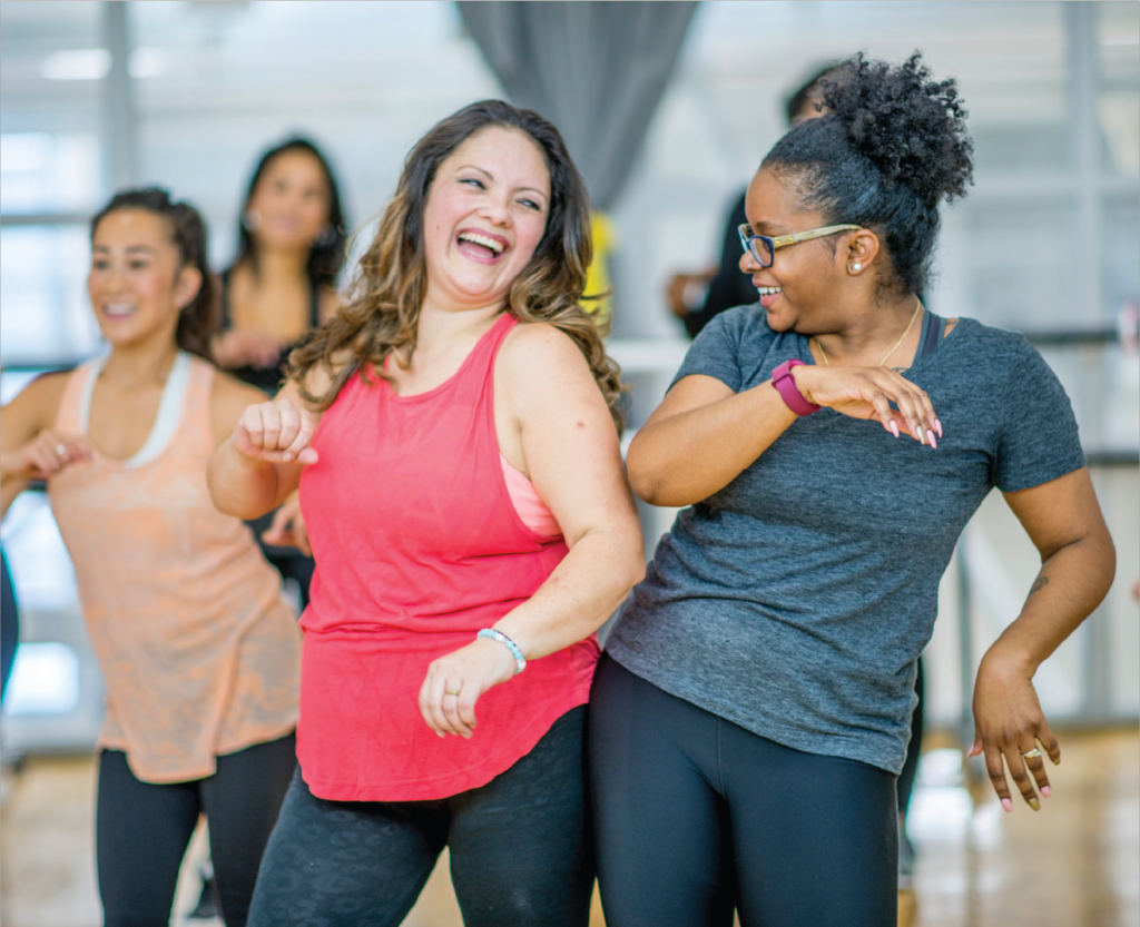 Two women bump their hips together while laughing in a dance class.