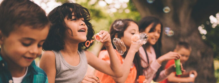 Five children stand in a diagonal line blowing bubbles into the sky