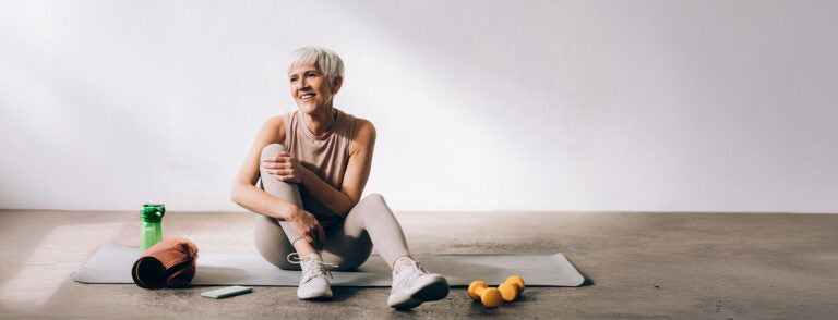 An older woman with short gray hair sits on a yoga mat with weights and kettlebells around her